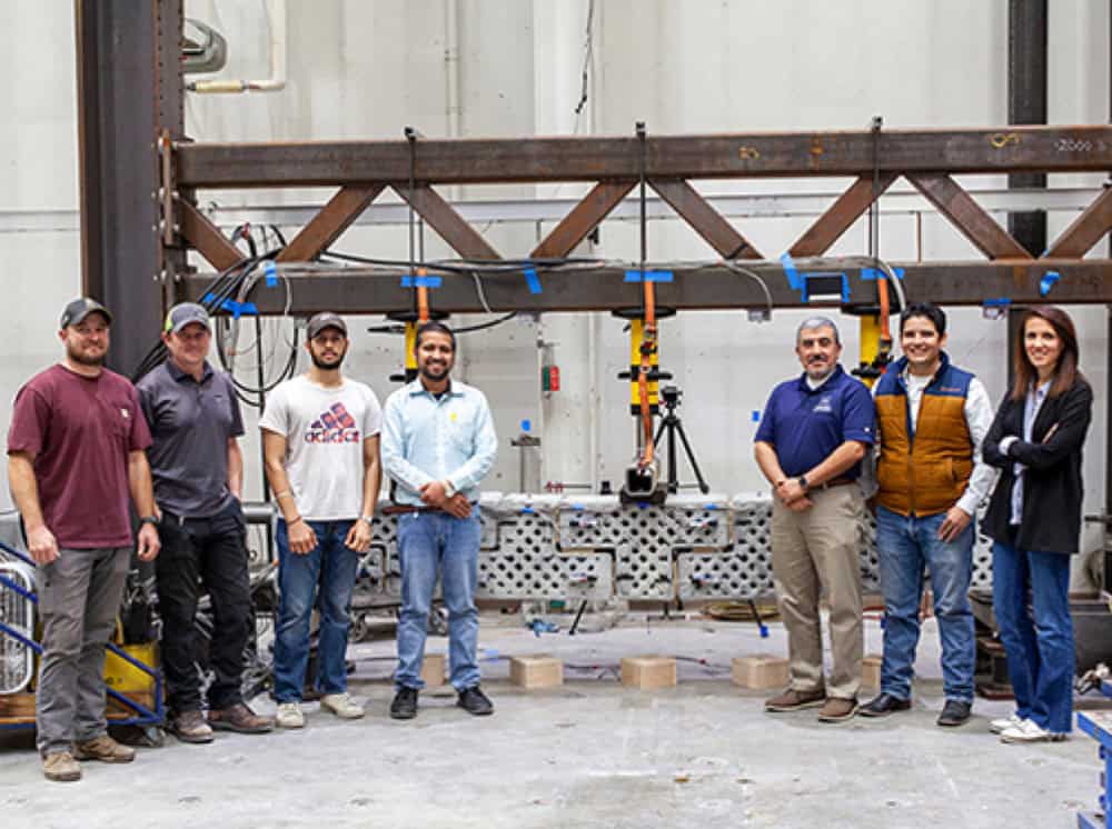 Eight people standing in a warehouse, posing in front of a large metal structure. The group appears to be a team, wearing casual clothing.