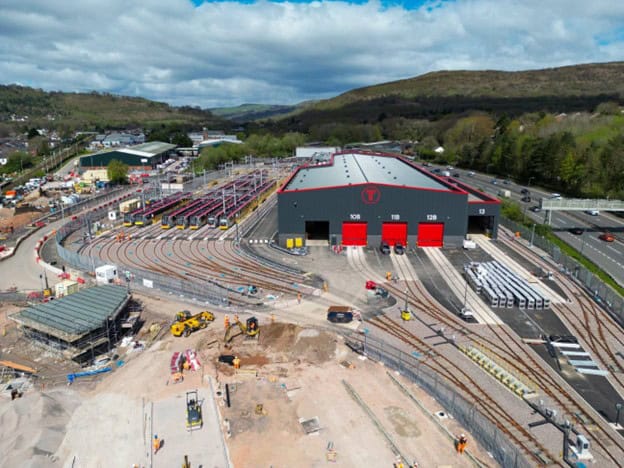 Aerial view of a rail depot with multiple train tracks, maintenance buildings, construction equipment, and surrounding greenery under a partly cloudy sky.