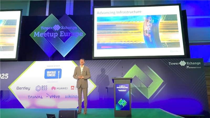 A man in a suit stands on stage giving a presentation titled "Advancing Infrastructure" at the TowerXchange Meetup Europe event. Sponsor logos are displayed on the stage background.