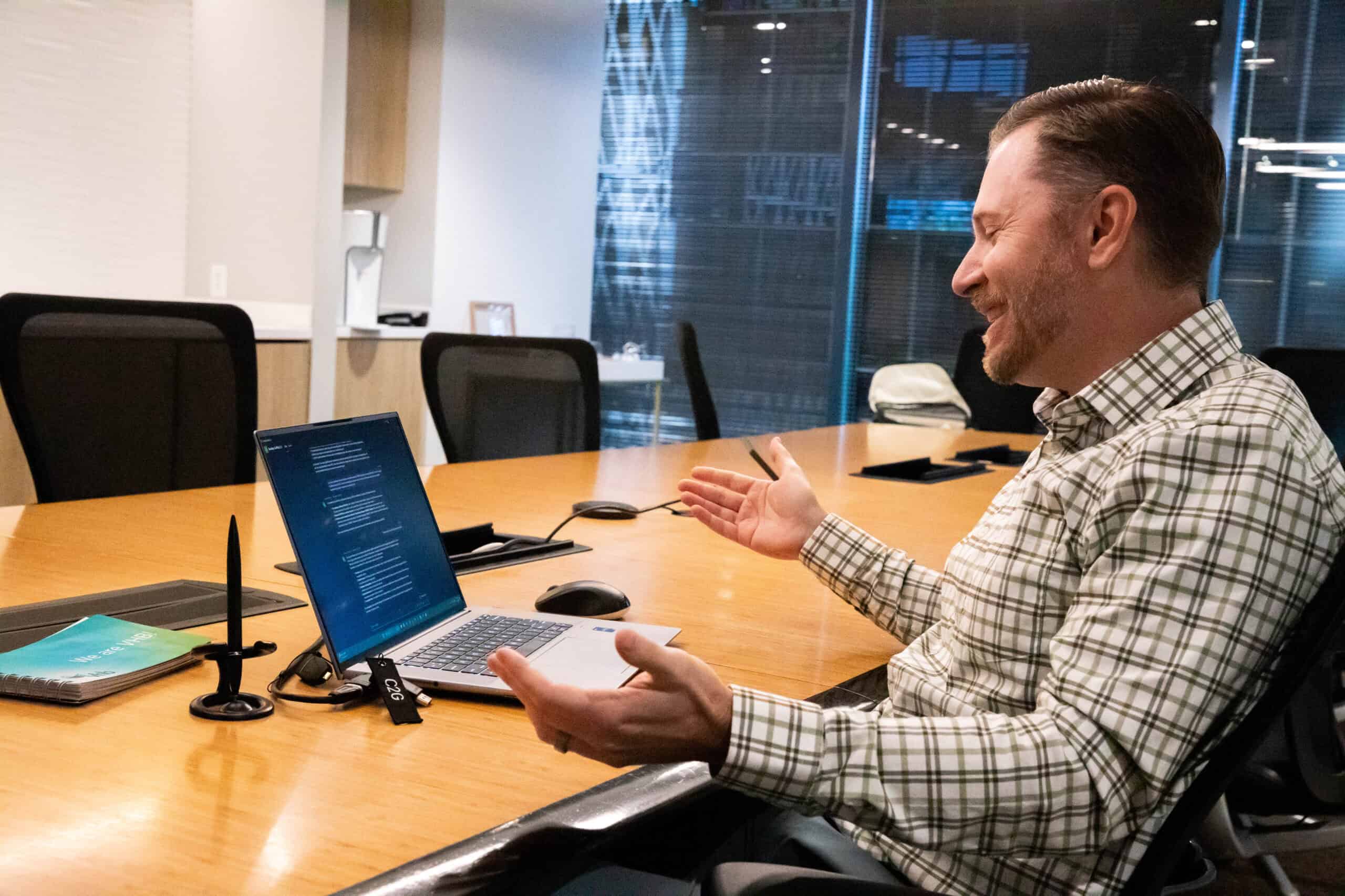 A man sits at a conference table, gesturing and speaking during a video call on his laptop in a modern office setting.