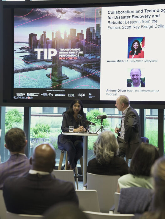 A woman and a man speak on stage at a conference, with a large screen behind them displaying event and speaker information about disaster recovery and the Key Bridge collapse.