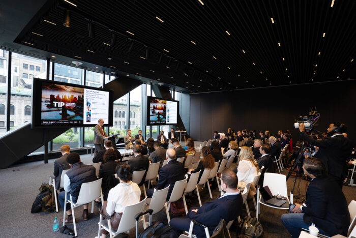 A group of people attends a panel discussion in a modern conference room with large windows, multiple screens, and a camera crew recording the event.
