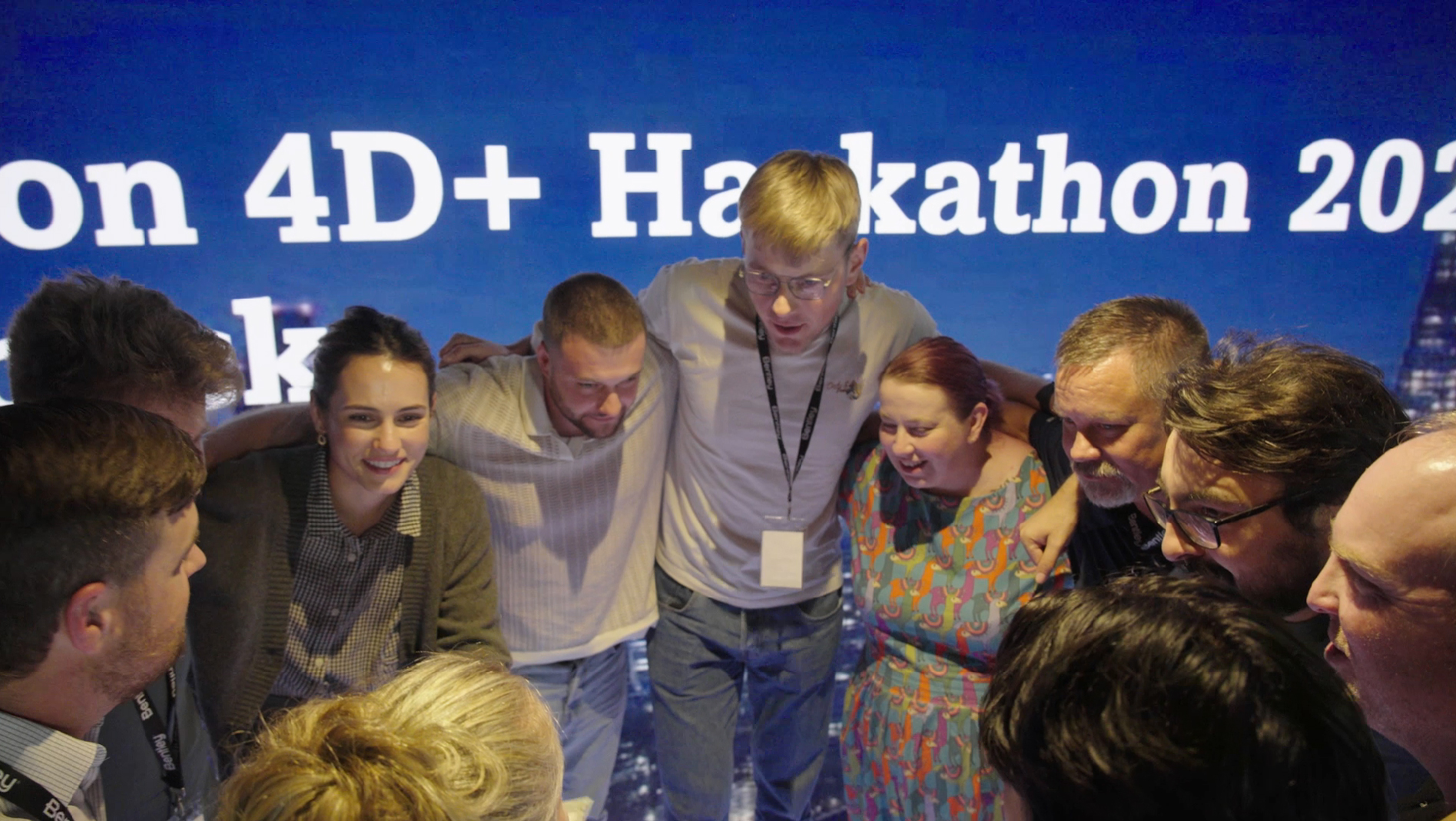 A group of people stands in a close circle with arms around each other, smiling and focusing inward, in front of a bright screen displaying "Hackathon 201" at a London event focused on construction and infrastructure innovation.