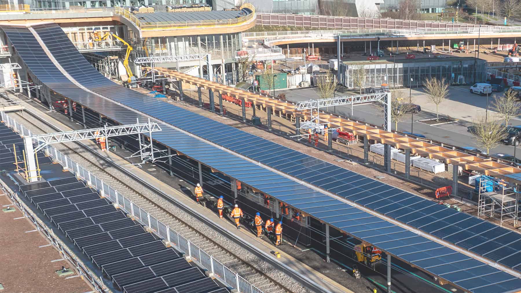A group of construction workers wearing orange vests walk along train tracks beneath large arrays of solar panels at a modern transportation facility under construction, reflecting the engineering clarity championed by the Arcadis Rail Division.