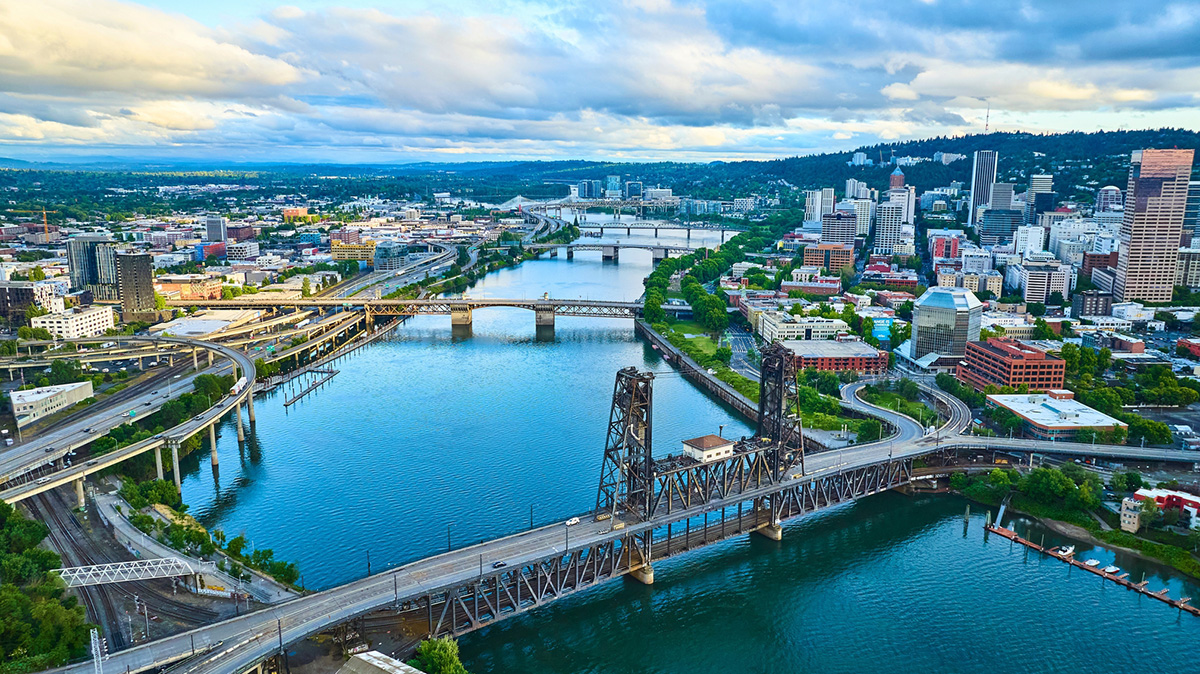 Aerial view of multiple bridges spanning a river in Portland, with high-rise buildings, green spaces, and hills in the background under a cloudy sky—showcasing sustainable city infrastructure.