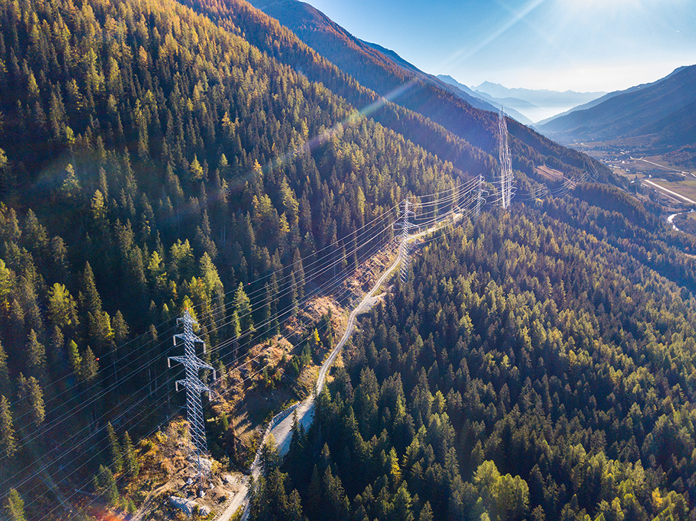 Aerial view of overhead power lines running through a forested mountain landscape under a clear sky, showcasing impressive power line engineering with distant valleys visible.
