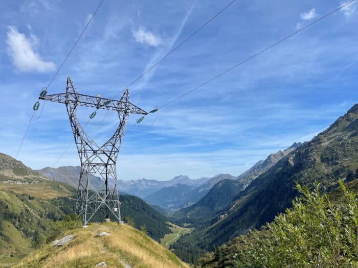 A metal electricity pylon stands on a grassy hill, with power lines stretching across a mountain valley under a blue sky with scattered clouds.