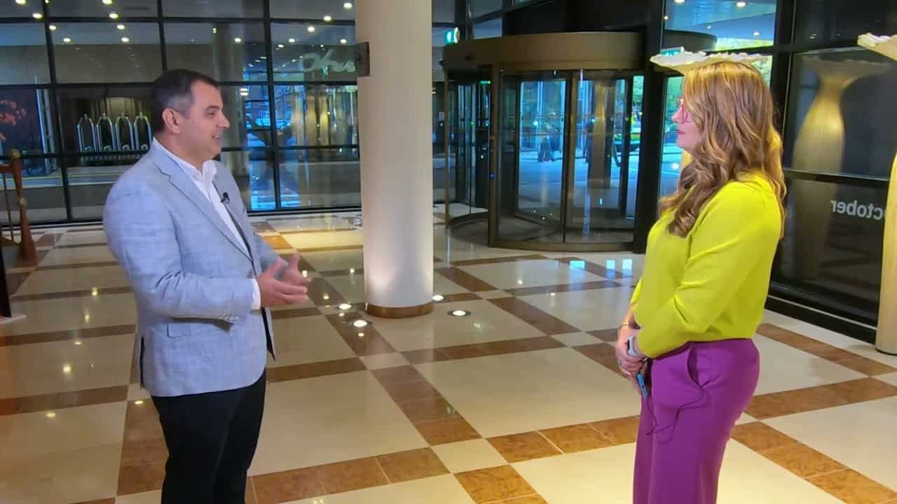 A man and a woman stand facing each other, discussing transportation value in a spacious, modern lobby with large windows and revolving doors.