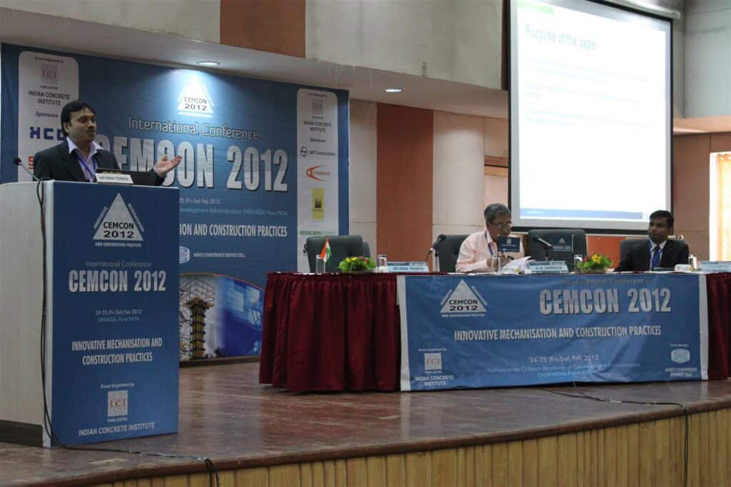 Apurba Tribedi presents at a podium during the CEMCON 2012 conference, discussing engineering advancements as two panelists sit at a table and a presentation is displayed on a screen in the background.