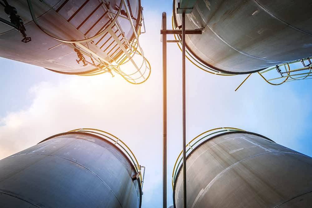 Four large industrial storage tanks viewed from below against a bright sky, with ladders and railings visible on the tanks' sides.