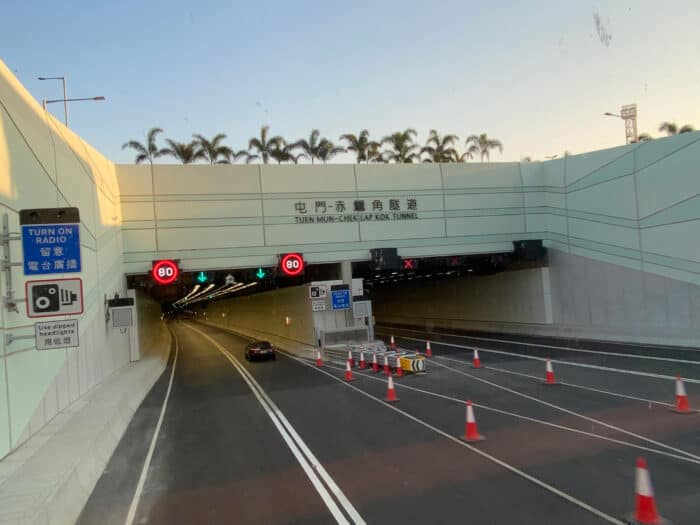 Entrance to the Tuen Mun-Chek Lap Kok Tunnel with traffic cones, speed limit signs, and vehicles entering the tunnel under clear sky.