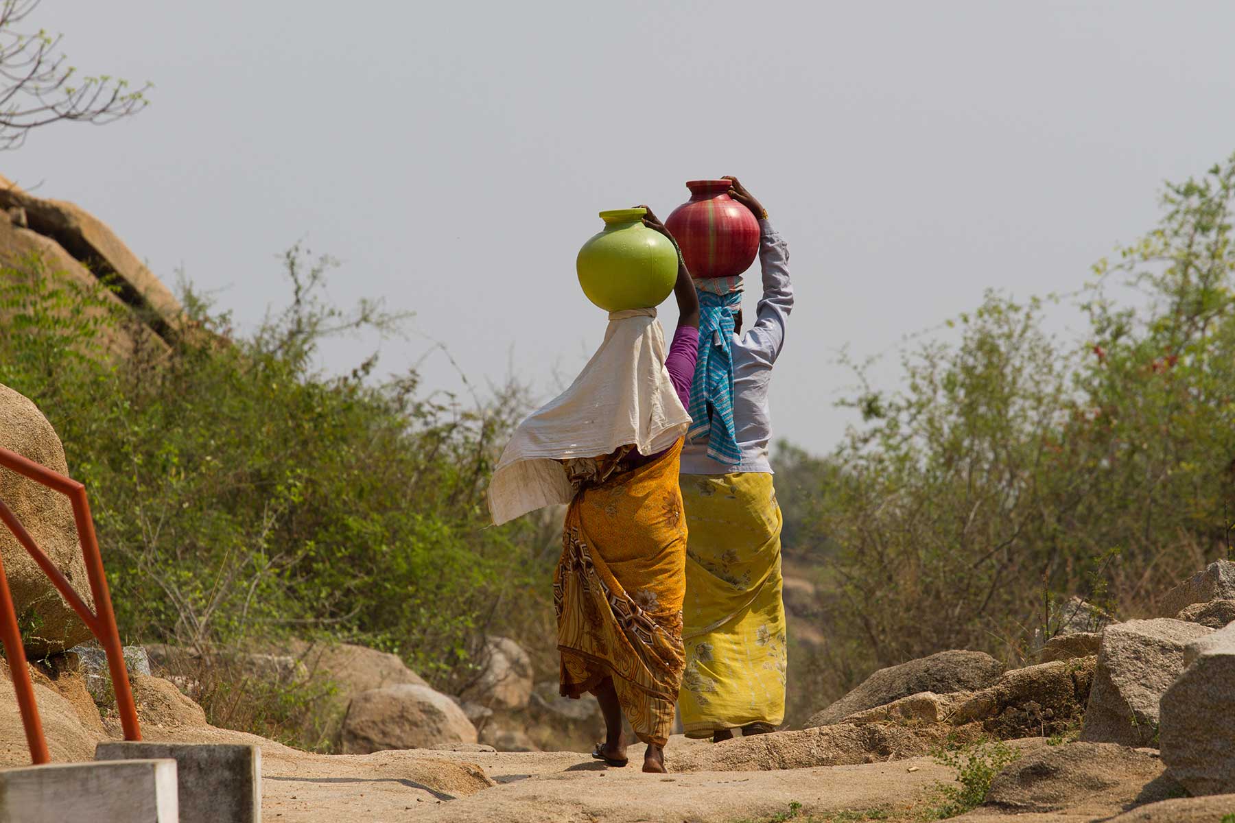 Two women walk on a rocky path carrying colorful water pots on their heads, while the natural background highlights how Digital Transformation can create solutions for remote communities.