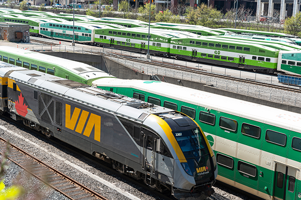 A VIA Rail passenger train and several green GO Transit commuter trains are parked on parallel tracks in a rail yard, reflecting Toronto rail protection efforts and advancements in rail safety innovation.