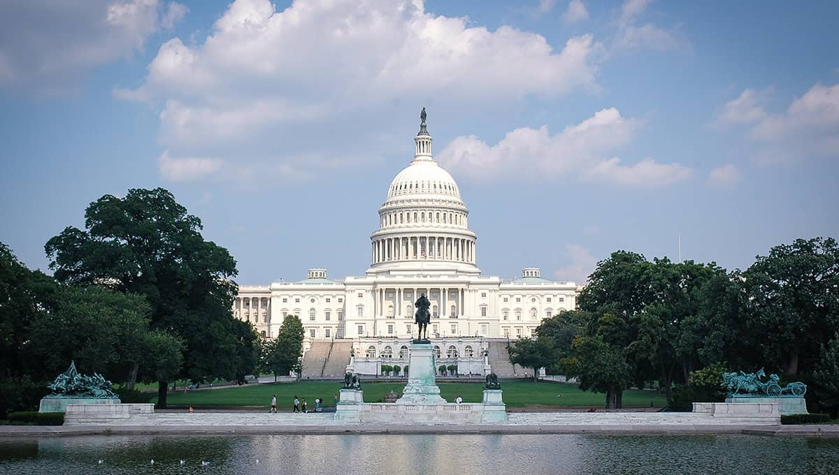 A view of the U.S. Capitol building in Washington, D.C., with a foreground of trees, statues, and a reflecting pool under a partly cloudy sky.