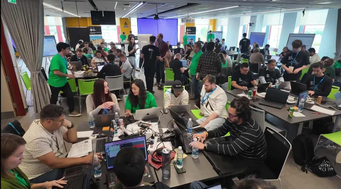 Large group of people working on laptops and collaborating at tables in a brightly lit, modern office or event space.