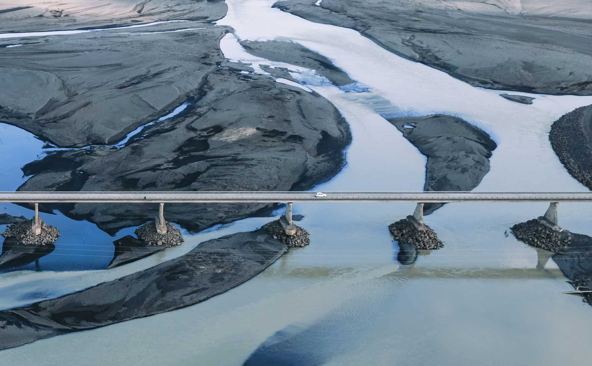 Aerial view of a bridge crossing a river with multiple channels and dark sediment, with a single car driving across.