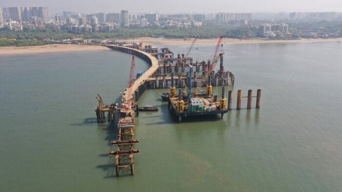 Aerial view of a bridge under construction over water, with cranes, machinery, and city buildings visible in the background.