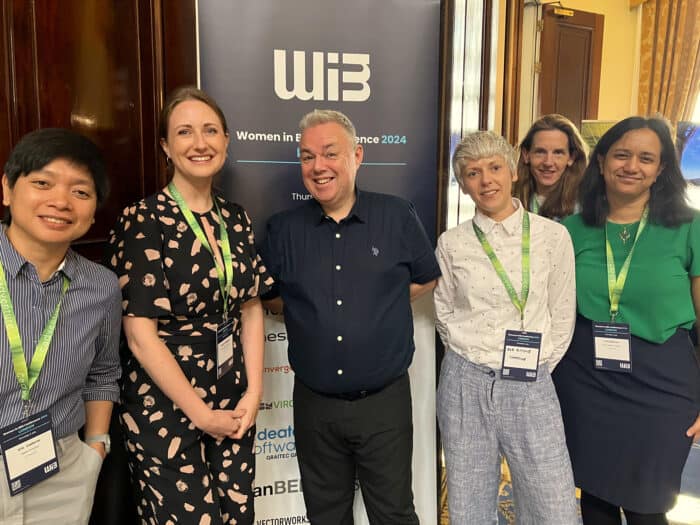 Group of six people smiling at a conference, standing in front of a "Women in Business Conference 2024" sign. They wear name badges and business-casual attire.