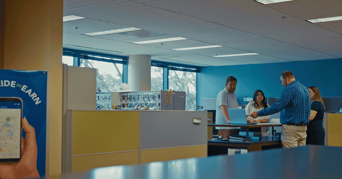 Four people stand and talk around a tall desk in a modern office with cubicles and large windows. A poster with a phone is visible on the left, highlighting the latest site design solutions like Bentley OpenSite+.