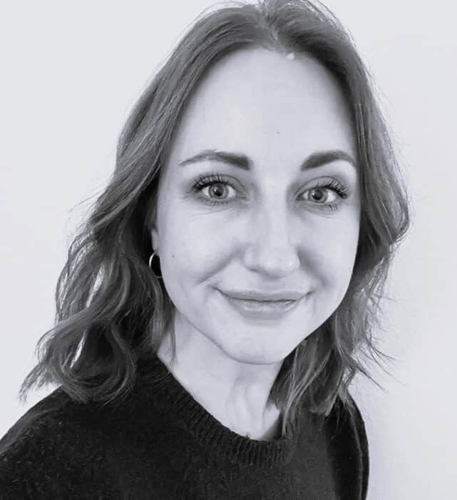 Black and white photo of a smiling person with wavy hair, wearing a dark top and hoop earrings, looking directly at the camera.