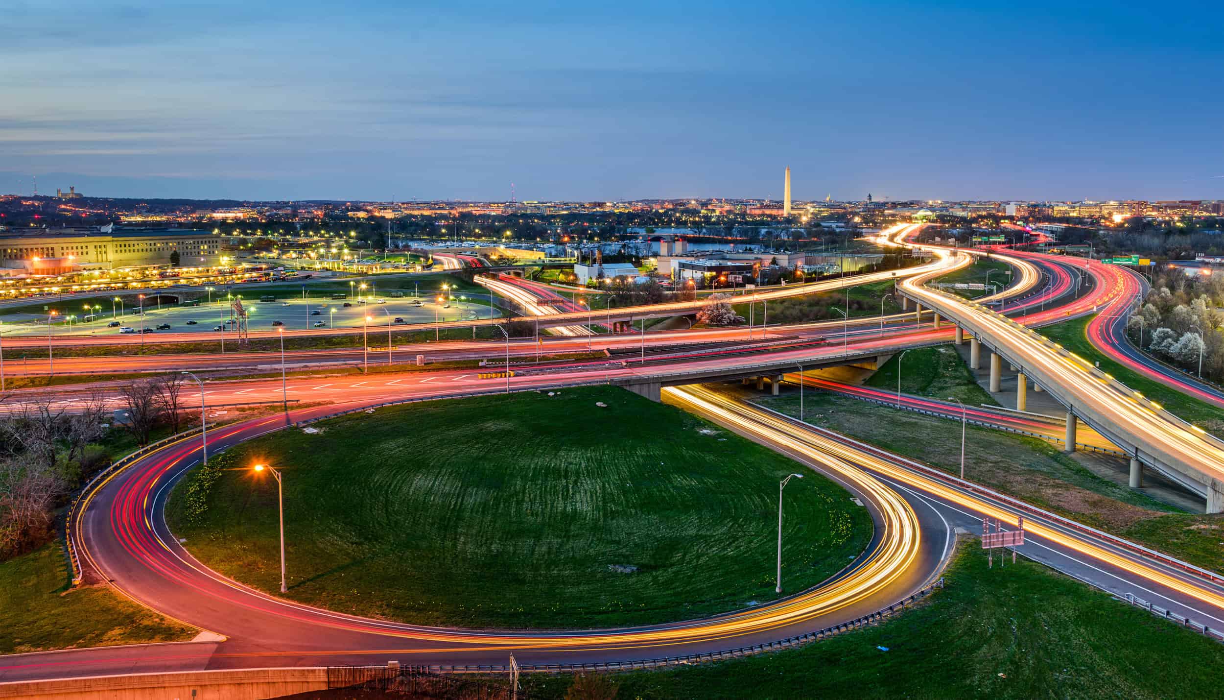 Aerial view of a busy highway interchange at dusk, with light trails from vehicles and a cityscape in the background.