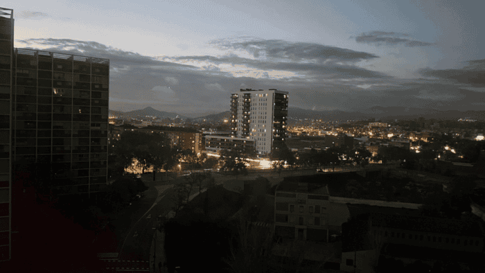 Cityscape at dusk with illuminated buildings, a central high-rise, dark tree silhouettes, and mountains in the background under a cloudy sky—the scene whispers stories of when the lights went out during the Iberian Blackout.