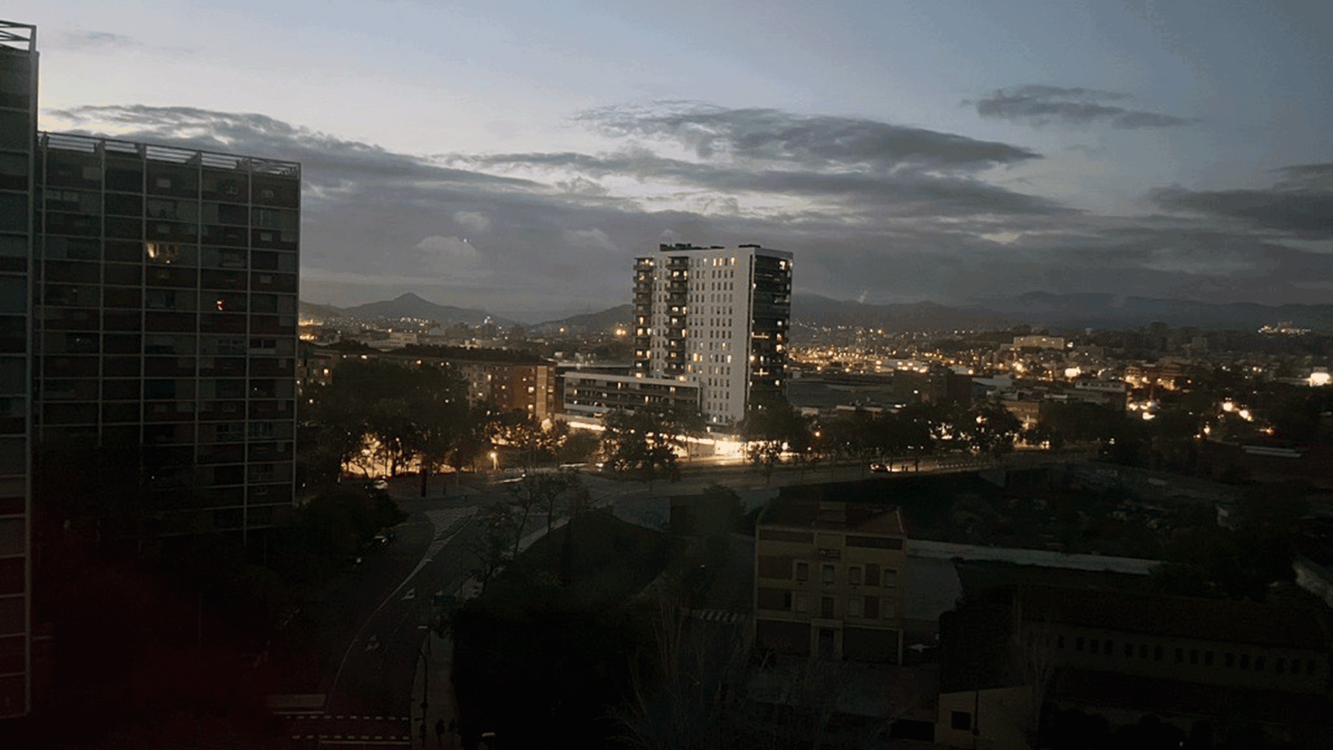 Cityscape at dusk with illuminated buildings, a central high-rise, dark tree silhouettes, and mountains in the background under a cloudy sky—the scene whispers stories of when the lights went out during the Iberian Blackout.