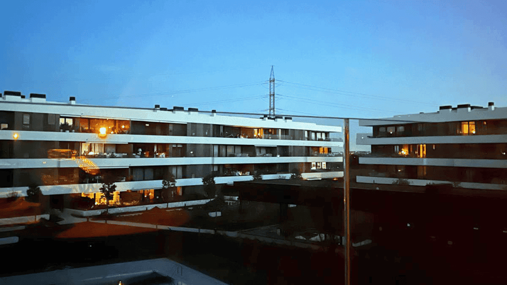View of modern apartment buildings at dusk, their illuminated windows hinting at Stories inside, while a power line tower stands in the background—a scene echoing memories of the Iberian Blackout when all the lights went out.