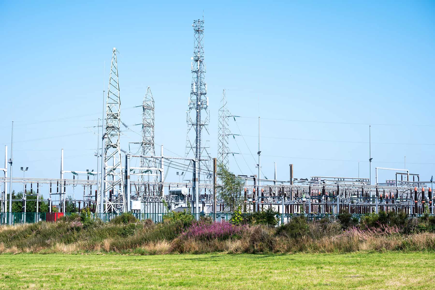 Electric power substation with multiple metal frameworks, transmission towers, and electrical equipment under a clear blue sky, surrounded by grass—ideal for analyzing or mapping a substation data model.