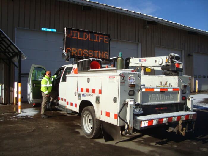 A worker standing beside a utility truck with "WILDLIFE CROSSING" displayed on an electronic sign. The truck is parked outside a large garage.