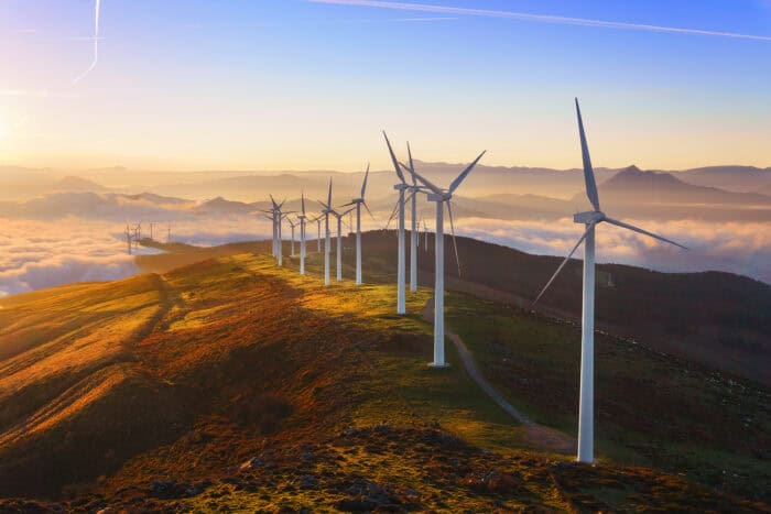 A row of wind turbines stands on a ridge at sunrise, with rolling hills and low clouds in the background.