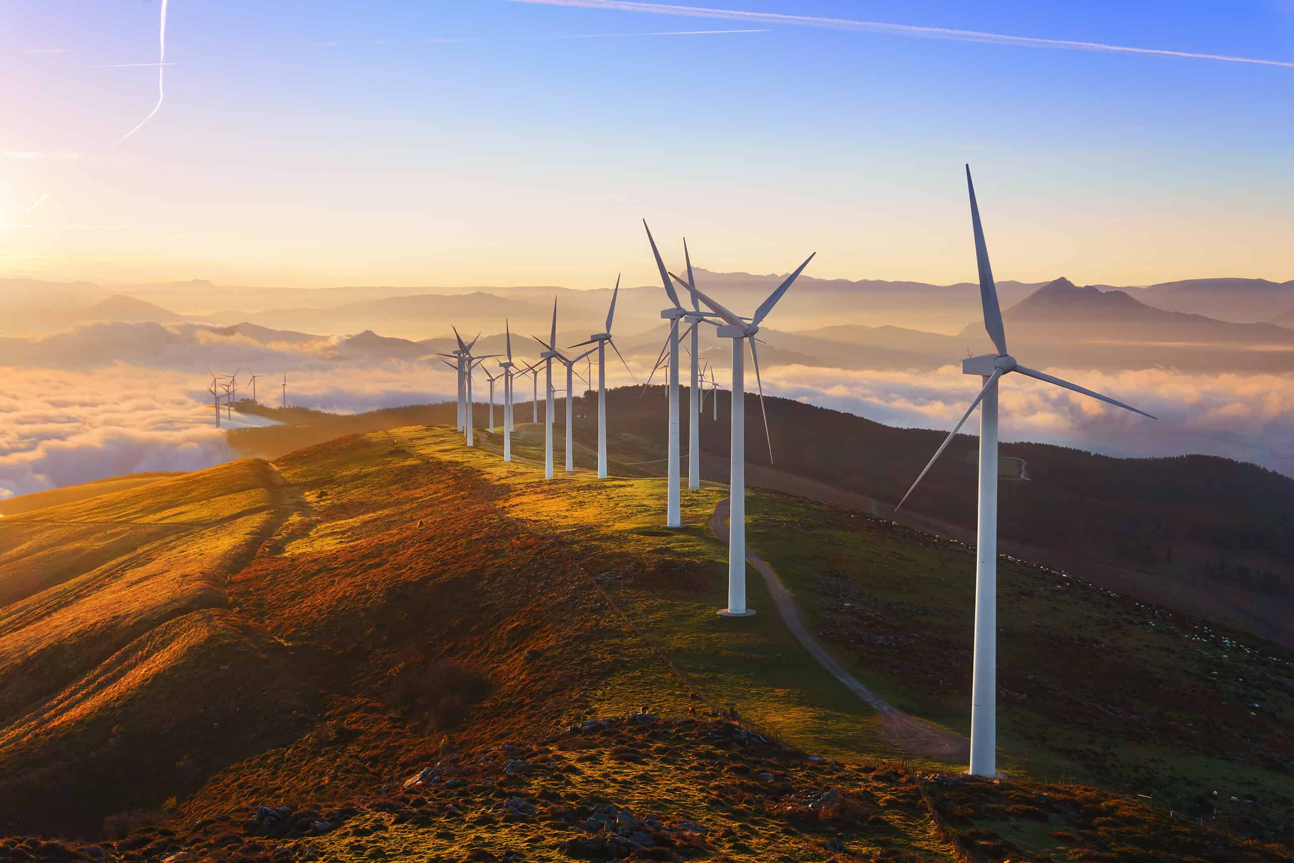 A row of wind turbines stands on a ridge at sunrise, with rolling hills and low clouds in the background.
