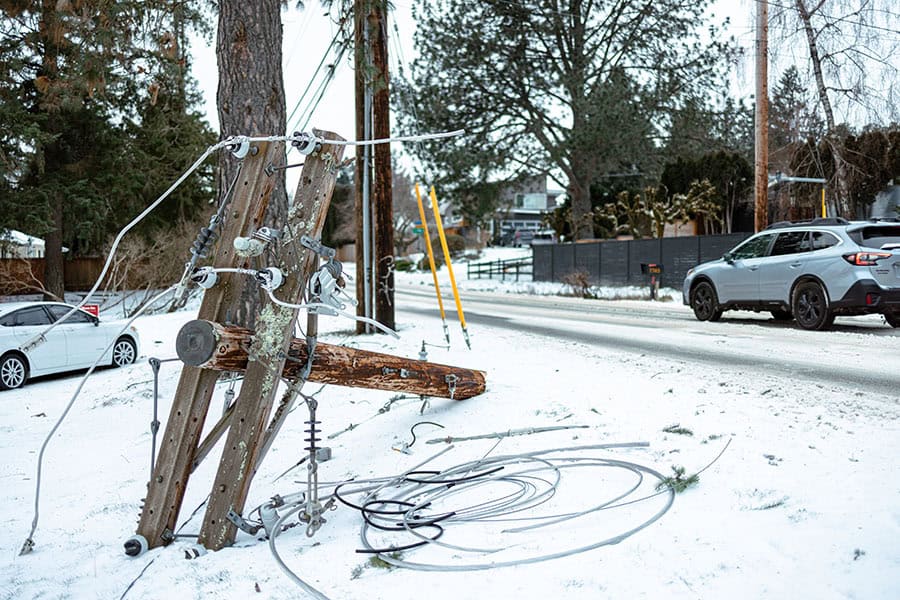 A broken utility pole with downed wires lies on snowy ground beside a road, with cars parked nearby and trees in the background.