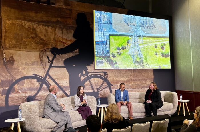 Four people sit on a stage panel discussing, with a large screen behind them displaying an aerial view of power lines—hinting at next-gen grid innovation—and a cyclist silhouette on the backdrop wall.