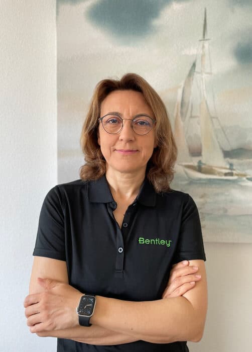 A woman with glasses and a black "Bentley" polo shirt stands with folded arms in front of a painting of a sailboat.