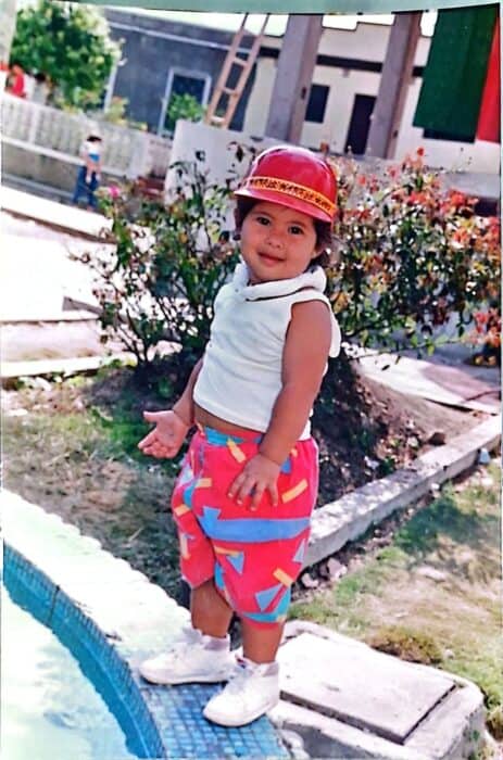 A young child in colorful shorts and a red hat stands by the edge of a pool, outdoors, with plants and buildings in the background.