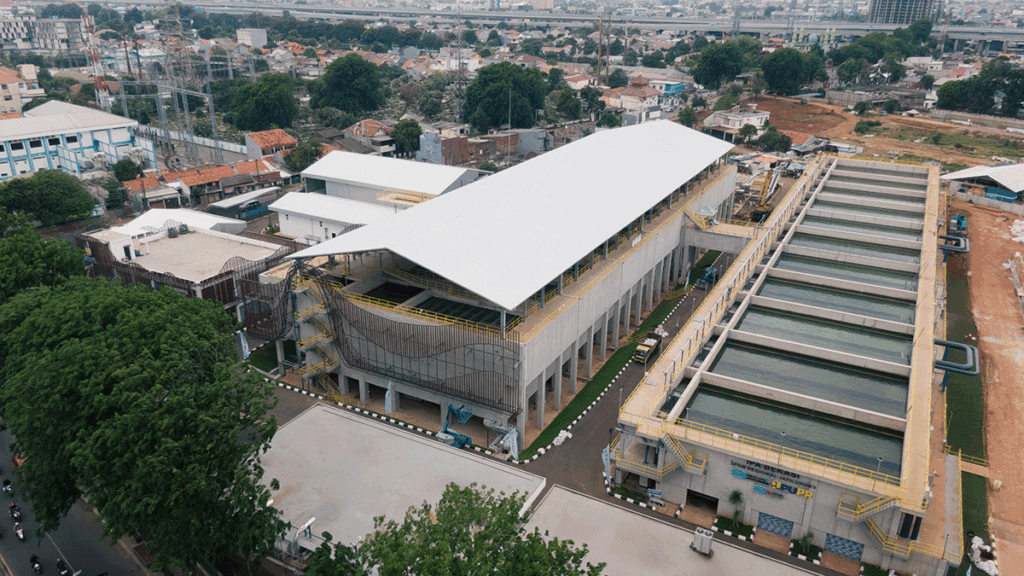 Aerial view of a large industrial facility with white roofing, multiple water tanks, and adjacent buildings in an urban area.