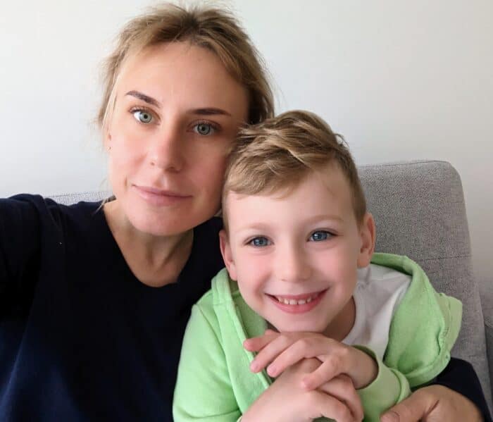 A woman and a young boy smile while sitting close together on a gray couch.