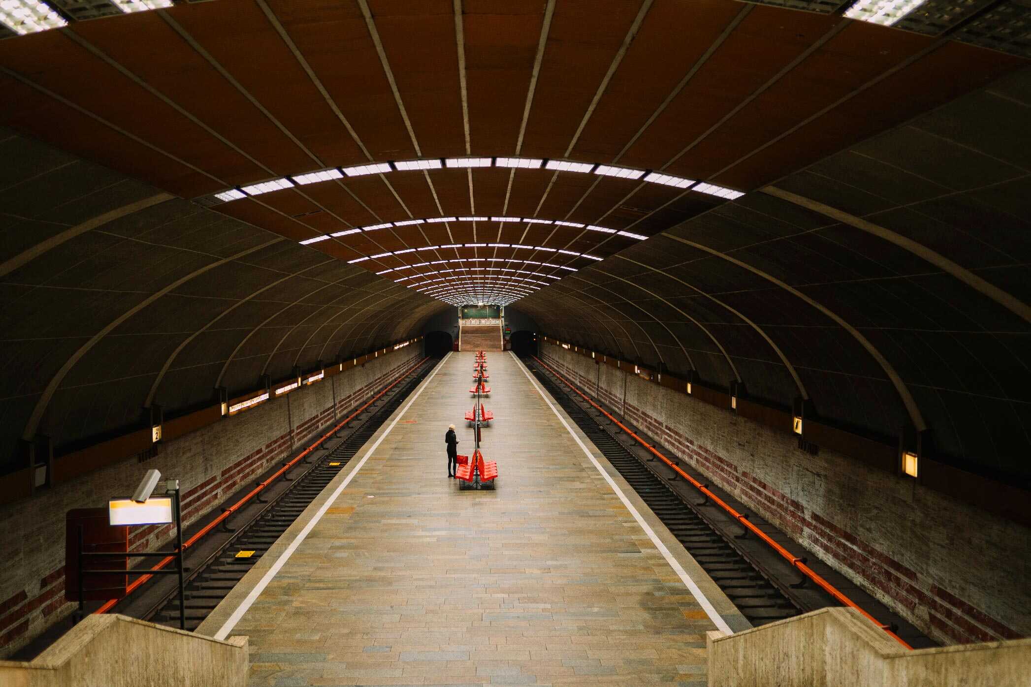 A person stands on an empty subway platform lined with orange cones, beneath a curved, well-lit ceiling, resembling an archive where moments are stored. No trains or signs of software-driven displays are present on the tracks.
