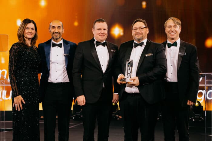 Five people in formal attire stand together on a stage, with one holding an award. A warm, glowing backdrop is behind them.