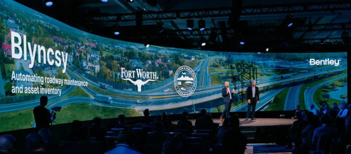 A conference stage with two presenters standing in front of a large screen displaying logos for Blyncsy, Fort Worth, and Bentley, with an image of a highway in the background.