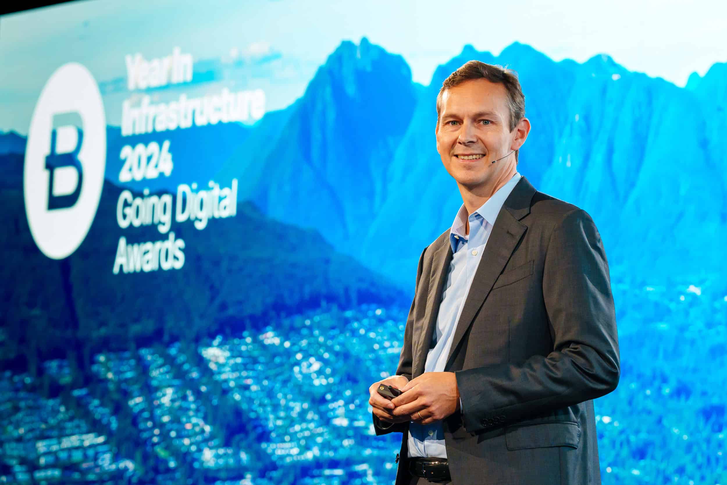 A man in a suit stands smiling in front of a screen displaying "Year in Infrastructure 2024 Going Digital Awards" with a mountain backdrop.