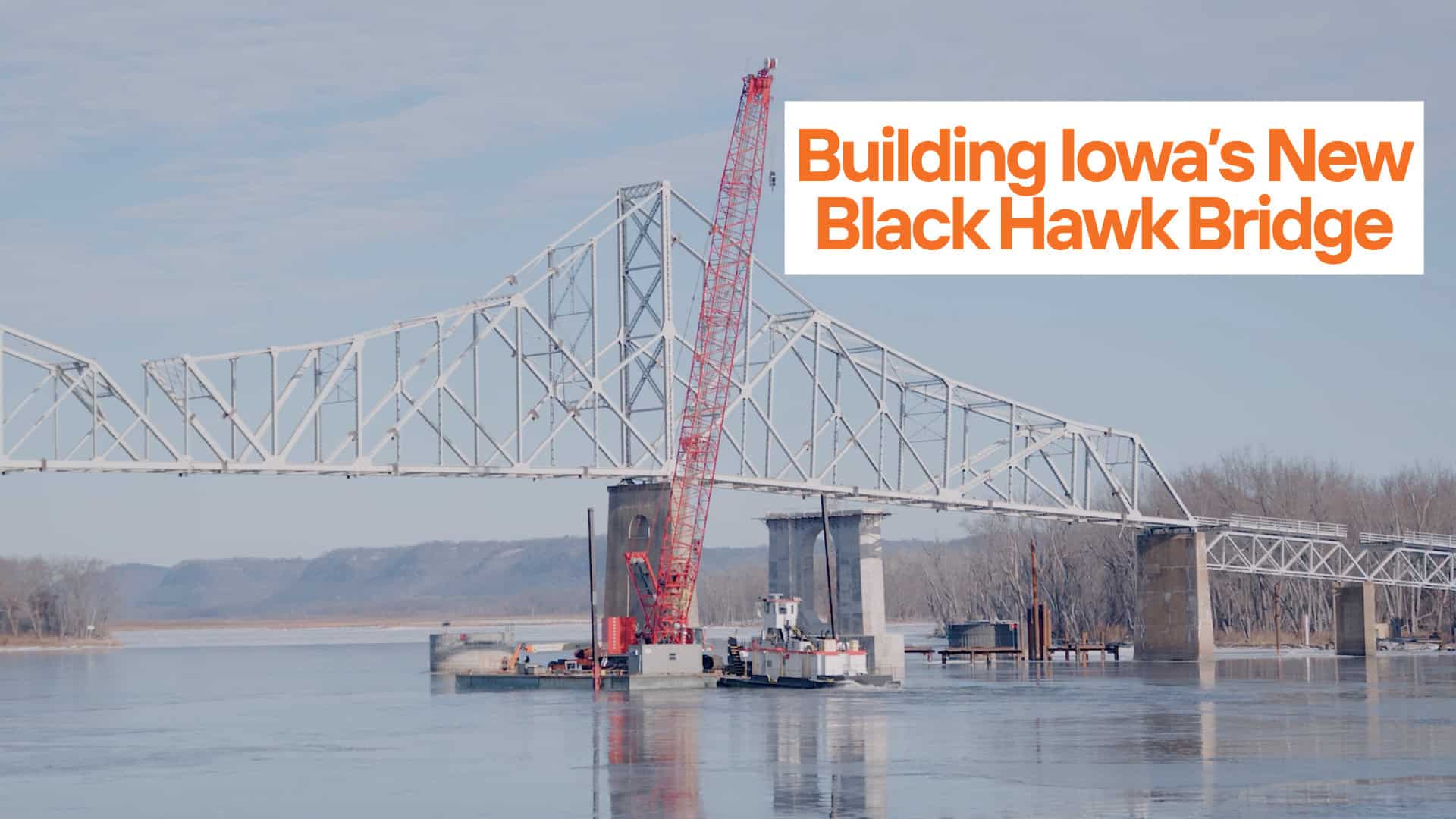 A construction crane and barge are positioned on the river at the partially built steel bridge, the Black Hawk Bridge in Iowa, under clear skies.
