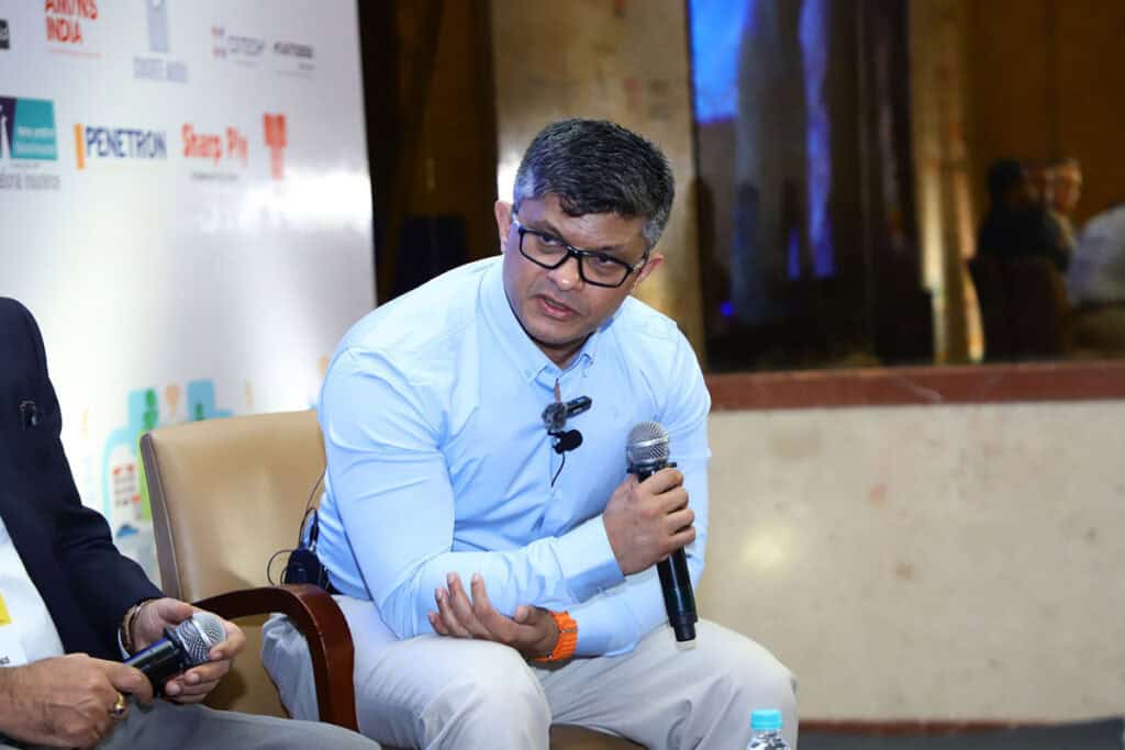 A man in glasses and a light blue shirt sits on a chair holding a microphone, appearing to speak at an indoor event or panel discussion on Geospatial Innovation hosted by WGIC and Bentley.