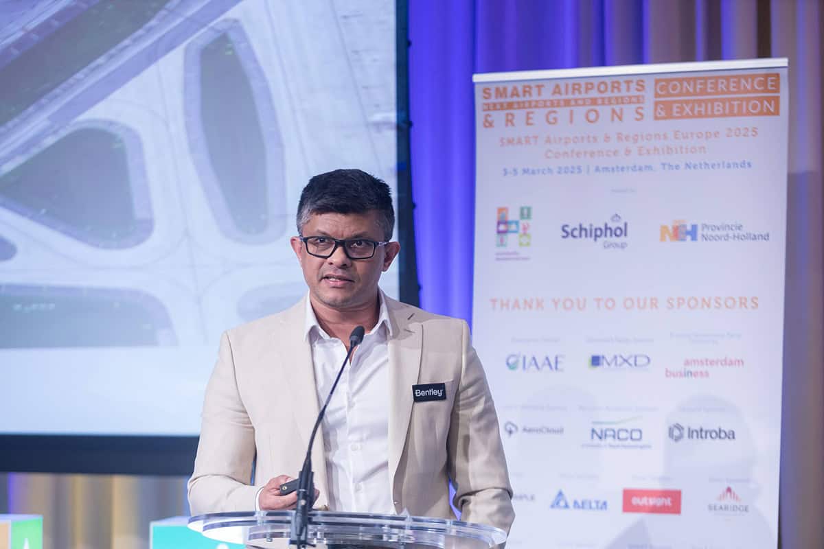 A man wearing glasses and a beige blazer with a "Bentley" name tag speaks about Geospatial Innovation at a podium during the SMART Airports & Regions Conference & Exhibition 2023.